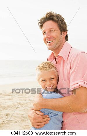 Stock Image - Father and son hugging on beach. Fotosearch