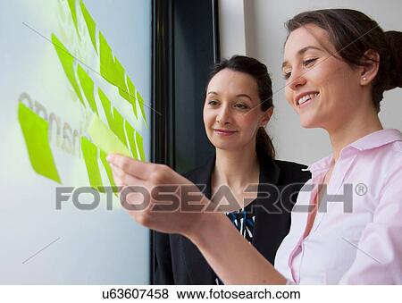 Stock Photo - Female colleagues sticking adhesive notes on glass. Fotosearch