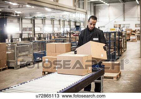Stock Image - Male warehouse worker checking cardboard box from conveyor belt. Fotosearch