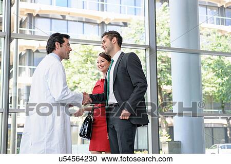Stock Image - Mature doctor shaking hands with young couple in hospital lobby. Fotosearch