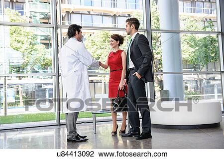 Mature doctor shaking hands with young couple in hospital lobby View Large Photo Image Stock Image - Mature doctor shaking hands with young couple in hospital lobby. Fotosearch