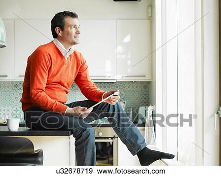 Stock Photo - Mature man holding tablet and sitting on kitchen worktop. Fotosearch