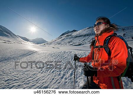 Stock Photograph - Mature woman hiking with backpack in sunlit snowy mountains, Skidadalur, Dalvik, Iceland. Fotosearch