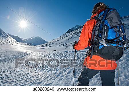 Stock Photograph - Mature woman hiking with backpack in sunlit snowy mountains, Skidadalur, Dalvik, Iceland. Fotosearch