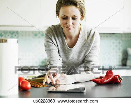 Stock Image - Mid adult woman using digital tablet on kitchen counter. Fotosearch