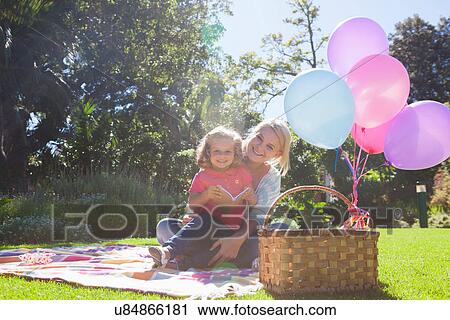 Stock Image - Mother and daughter sitting on blanket in garden with book and balloons. Fotosearch