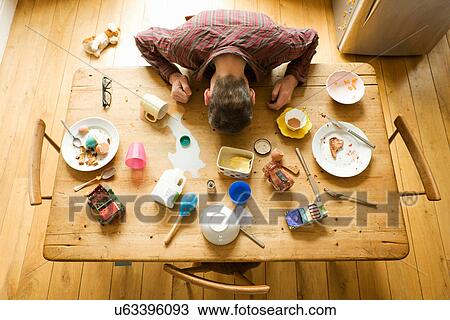 Stock Image - Overhead view of breakfast table with mature man amongst messy plates. Fotosearch