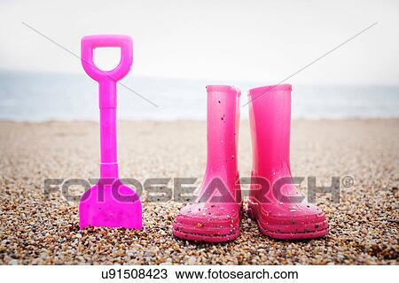 Stock Image - Pink spade and wellington books standing on beach. Fotosearch