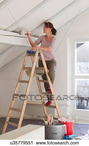 Stock Image - Woman on stepladders painting white ceiling. Fotosearch