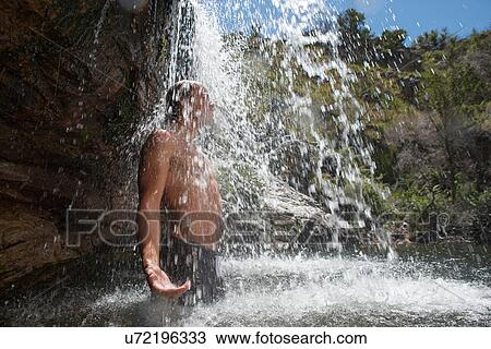 Stock Image - Young man standing under waterfall. Fotosearch