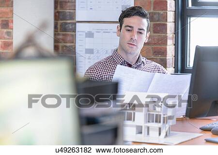 Stock Image - Architect inspecting plans at desk with model building. Fotosearch