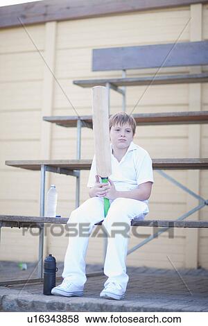 Stock Photo - Boy holding cricket bat sitting on bleachers. Fotosearch