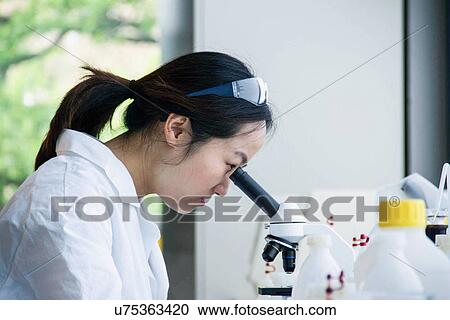 Stock Image - Close up of female scientist looking through microscope. Fotosearch