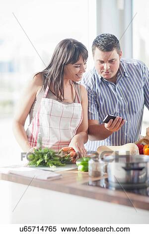 Stock Photography - Couple looking at smartphone whilst preparing food. Fotosearch