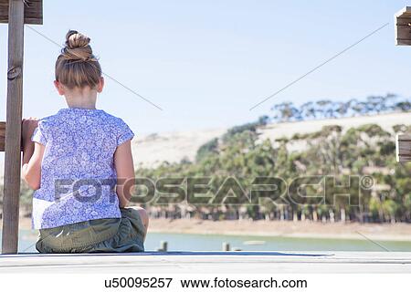 Stock Photo - Girl sitting on pier, rear view. Fotosearch