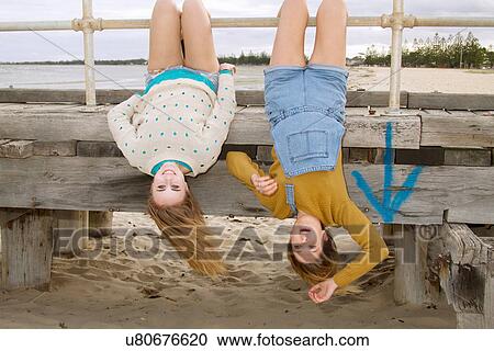Stock Image - Girlfriends hanging upside down from pier. Fotosearch