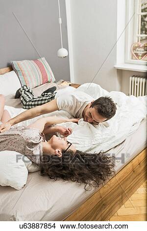 Picture - Mid adult couple wearing pyjamas lying on bed tickling. Fotosearch