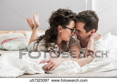 Stock Photo - Mid adult couple wearing pyjamas lying on front on bed, face to face. Fotosearch