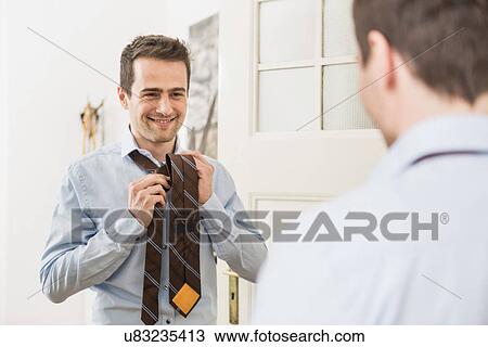 Stock Image - Mid adult man fastening tie in mirror. Fotosearch