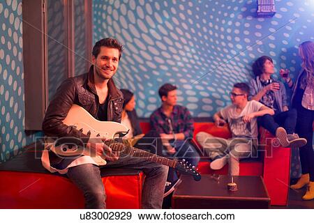 Stock Photo - Mid adult man holding guitar sitting in front of group of teenagers. Fotosearch