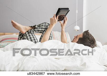 Mid adult man lying on bed using digital tablet View Large Photo Image Stock Photo - Mid adult man lying on bed using digital tablet. Fotosearch