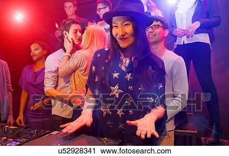 Stock Image - Mid adult woman DJing at party, people dancing in background. Fotosearch