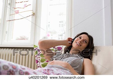 Stock Photo - Mid adult woman lying on sofa listening to music through headphones. Fotosearch