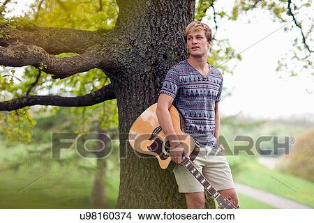 Picture - Portrait of young man leaning on tree trunk with guitar. Fotosearch