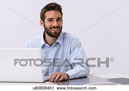 Stock Image - Portrait of young man sitting at desk with laptop. Fotosearch