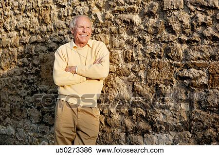 Stock Photograph - Senior man leaning against stone wall with arms crossed. Fotosearch