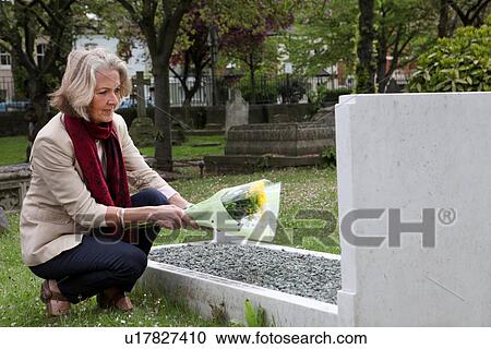 Stock Image - Senior woman holding flowers at grave stone. Fotosearch