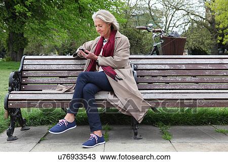 Stock Image - Senior woman sitting on park bench and looking at mobile phone. Fotosearch