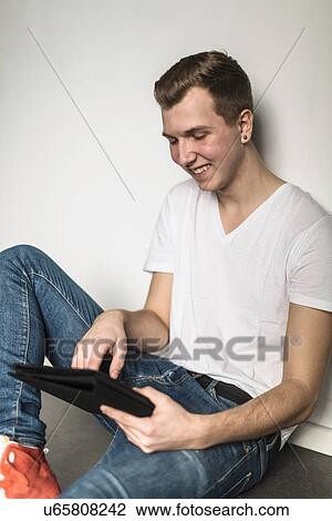 Stock Image - Teenage boy sitting on floor, using tablet. Fotosearch
