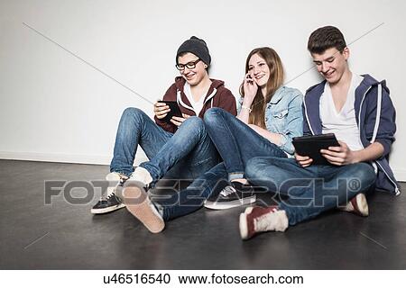 Stock Image - Three teenagers sitting on floor using technology. Fotosearch