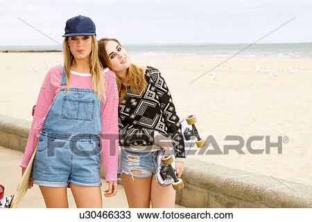 Stock Image - Two young women holding skateboards at beach. Fotosearch