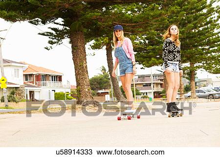 Stock Image - Two young women skateboarding through tree lined street. Fotosearch