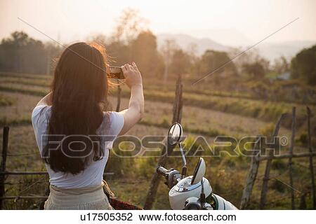 Stock Image - Woman on moped taking photograph in rural scene. Fotosearch