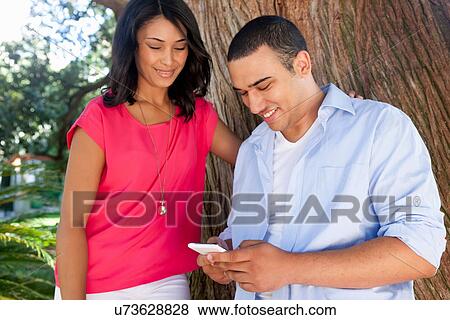 Young couple by tree trunk, man using smartphone View Large Photo Image Stock Photo - Young couple by tree trunk, man using smartphone. Fotosearch