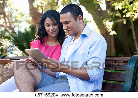 Young couple sitting on bench using digital tablet View Large Photo Image Picture - Young couple sitting on bench using digital tablet. Fotosearch