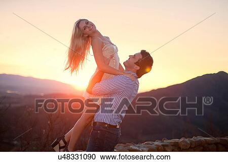 Stock Image - Young man lifting up girlfriend at sunset. Fotosearch