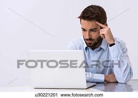 Young man sitting at desk looking at laptop View Large Photo Image Stock Image - Young man sitting at desk looking at laptop. Fotosearch