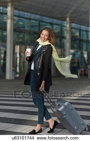 Picture - Young woman at airport with wheeled case and coffee. Fotosearch