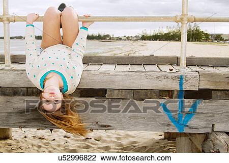 Stock Image - Young woman hanging upside down from pier. Fotosearch