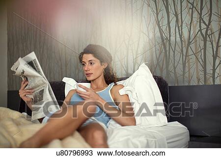 Stock Image - Young woman in bed with newspaper and coffee. Fotosearch