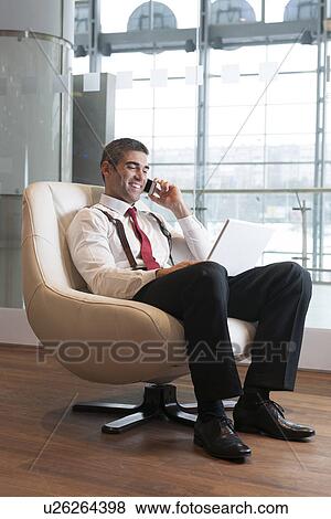 Stock Photo - Businessman in arm chair working on laptop on the phone. Fotosearch
