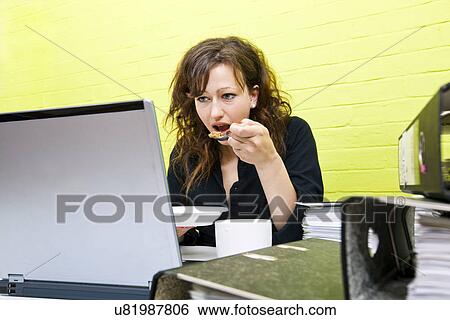 Stock Photograph - Caucasian young woman eating and working on her laptop computer at her desk. Fotosearch