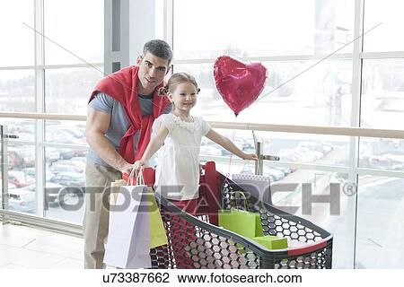 Stock Image - Father pushing young daughter in shopping trolley with shopping bags. Fotosearch