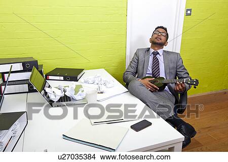 Indian businessman asleep at his desk clutching ukulele View Large Photo Image Picture - Indian businessman asleep at his desk clutching ukulele. Fotosearch