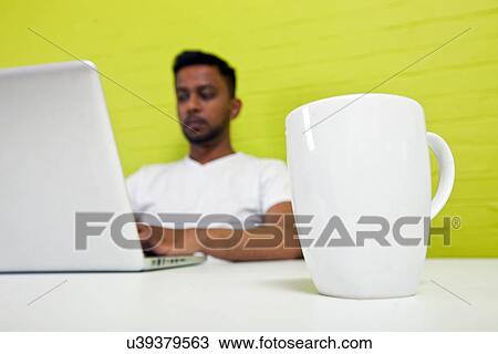 Stock Image - Indian man working at his desktop with mug in focus in foreground. Fotosearch