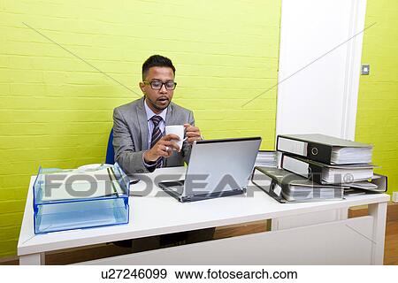Stock Photo - Portrait of Indian Businessman holding mug and working on his laptop computer at his desk. Fotosearch